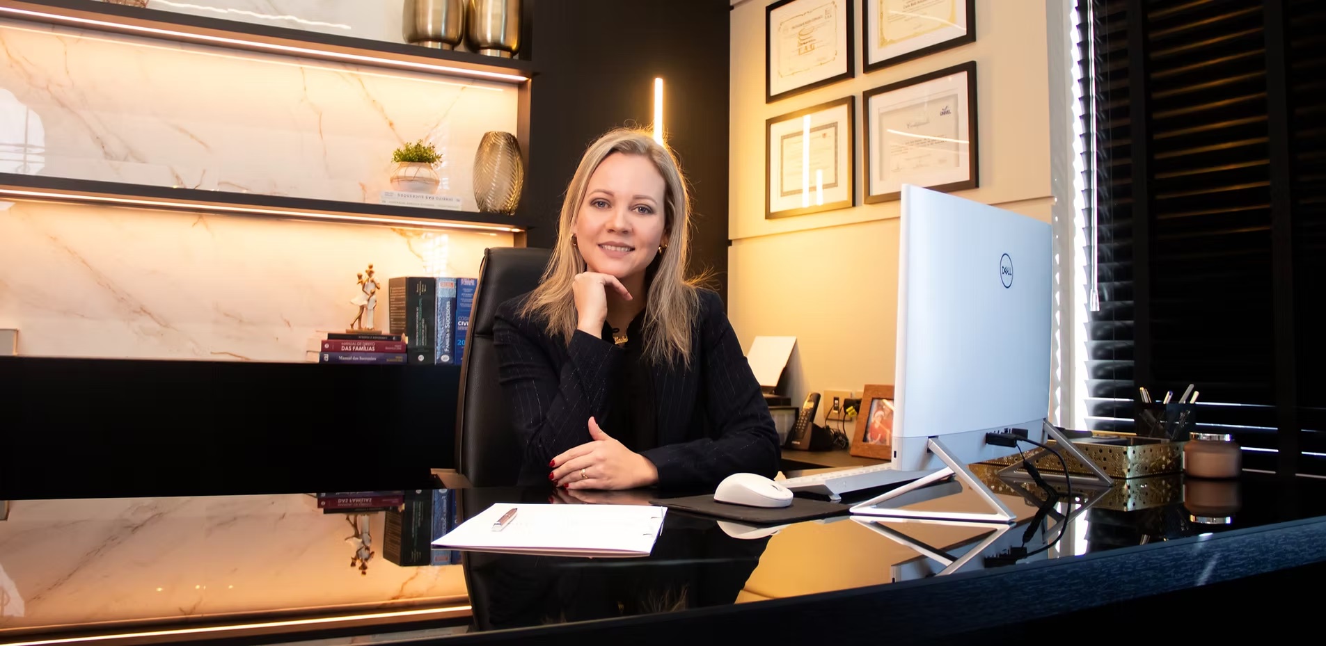 Carla Schons à mesa do escritório, com a estante iluminada e o monitor